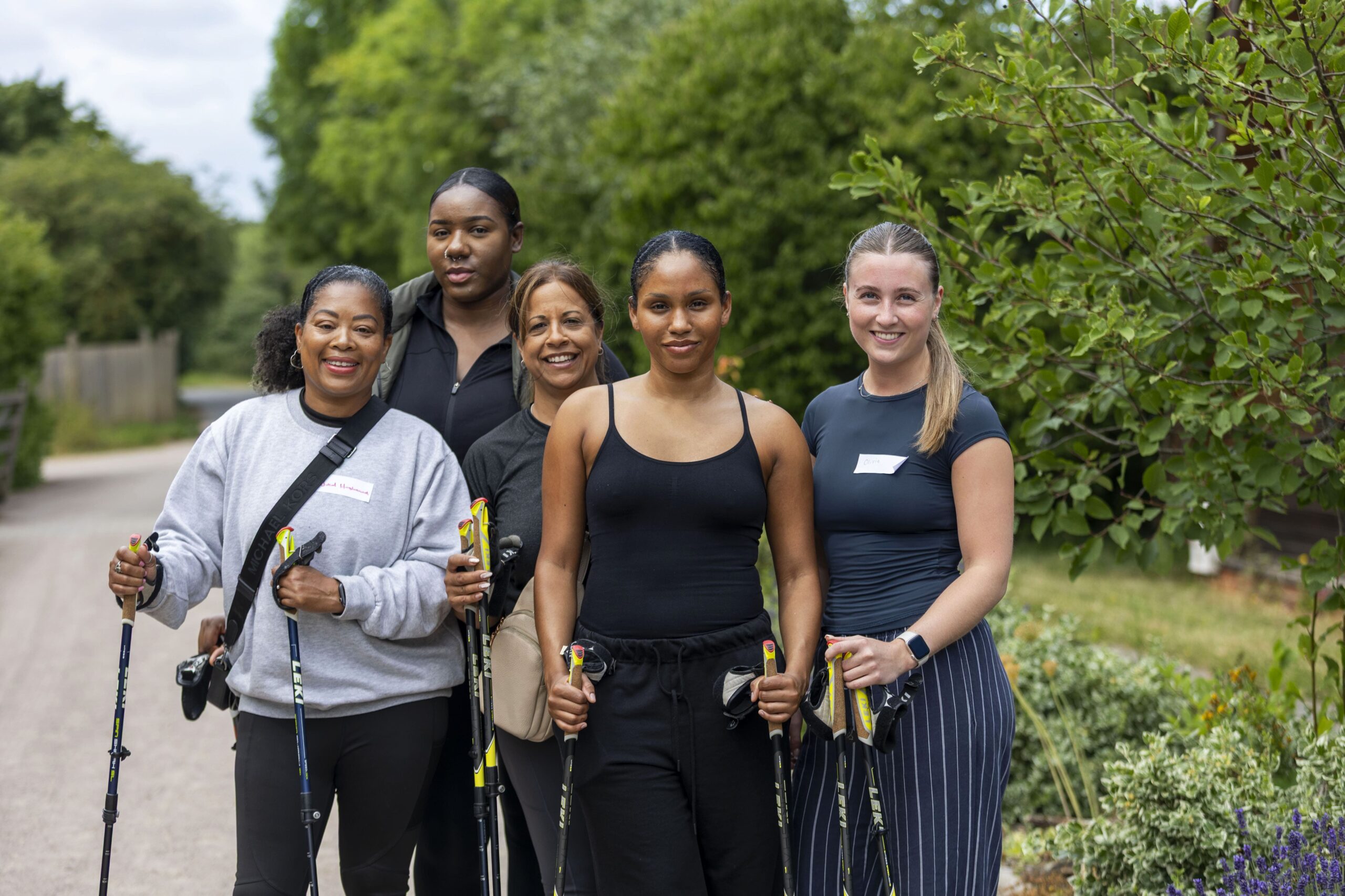 Five people with walking poles stand on a path outdoors, dressed for hiking, with trees and greenery in the background.
