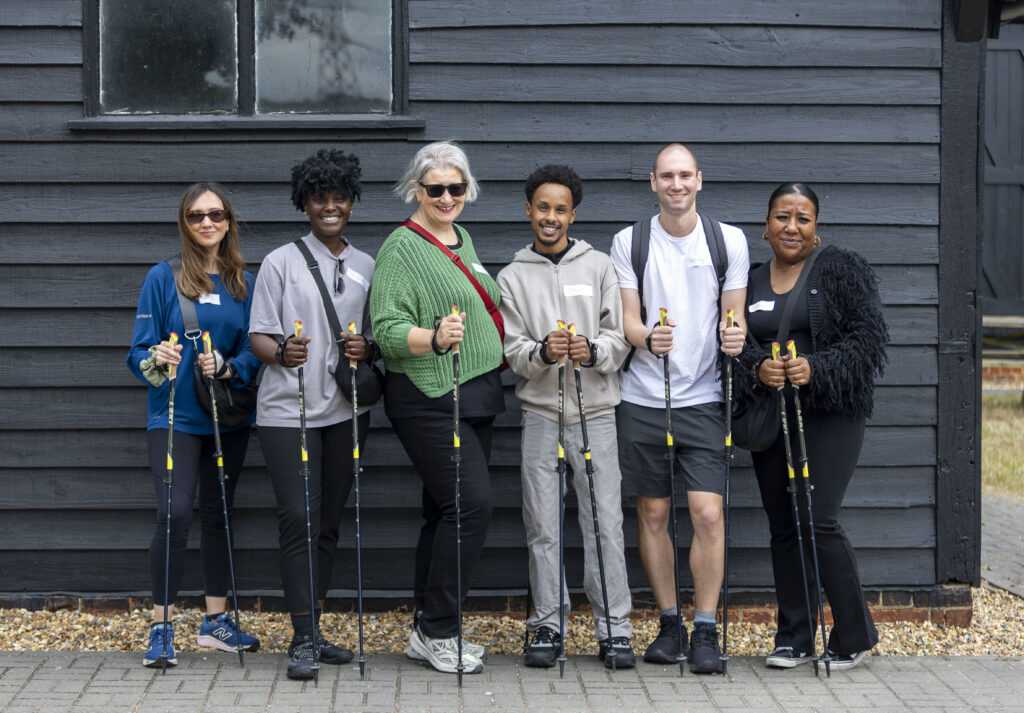 Six people with trekking poles stand in front of a dark wooden building, dressed for outdoor activity.