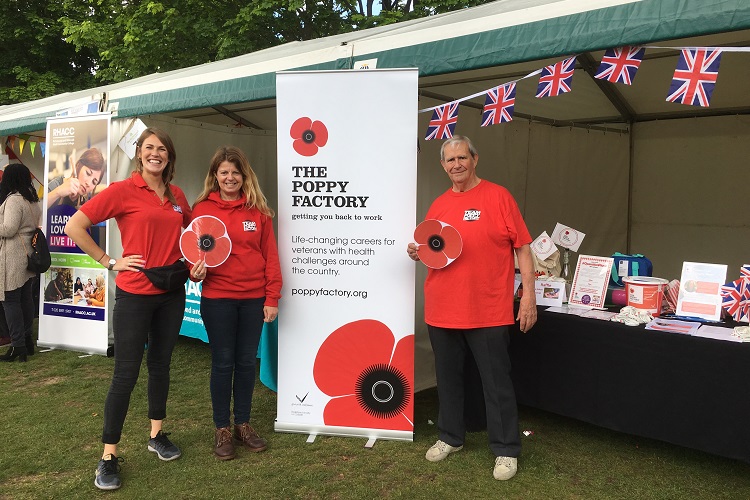 Three people in red shirts stand beside a Poppy Factory banner at an outdoor event.