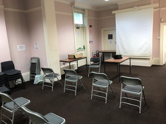 Small meeting room in Fairkytes with eight grey folding chairs facing a projection screen, two front tables with a box and projector, and light pink walls with posters.