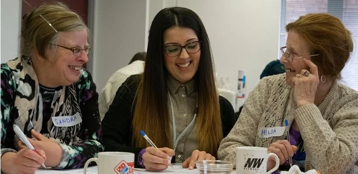 Three people seated at a table with mugs, papers, and pens; two wear name tags reading 'Sandra' and 'Hilda'.