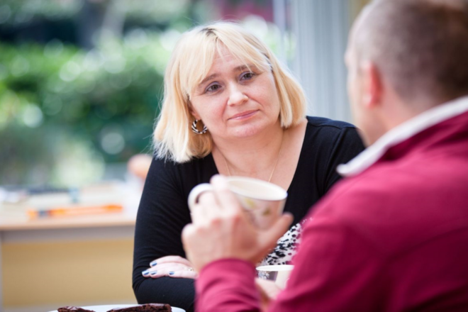 Two people sitting at a table indoors, conversing; one holds a cup.