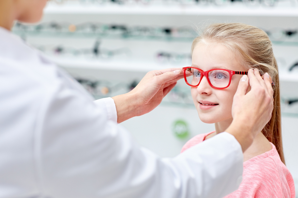 Optometrist fitting red eyeglasses on a young girl at an opticians, with shelves of eyeglasses in the background.