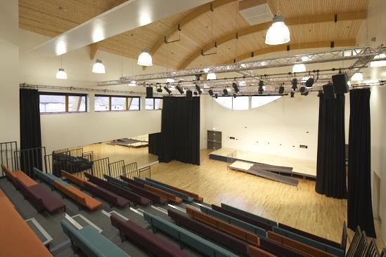 Empty auditorium with wooden floor, stage with black curtains, tiered seating with colourful cushions, arched wooden ceiling, and large windows.