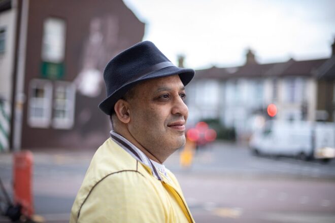 Person in yellow jacket and black hat standing on a city street with buildings, mural, and vehicles in the background.