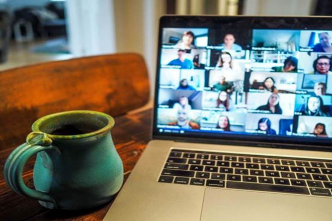 Laptop on wooden table showing a video conference, with a green coffee mug beside it.