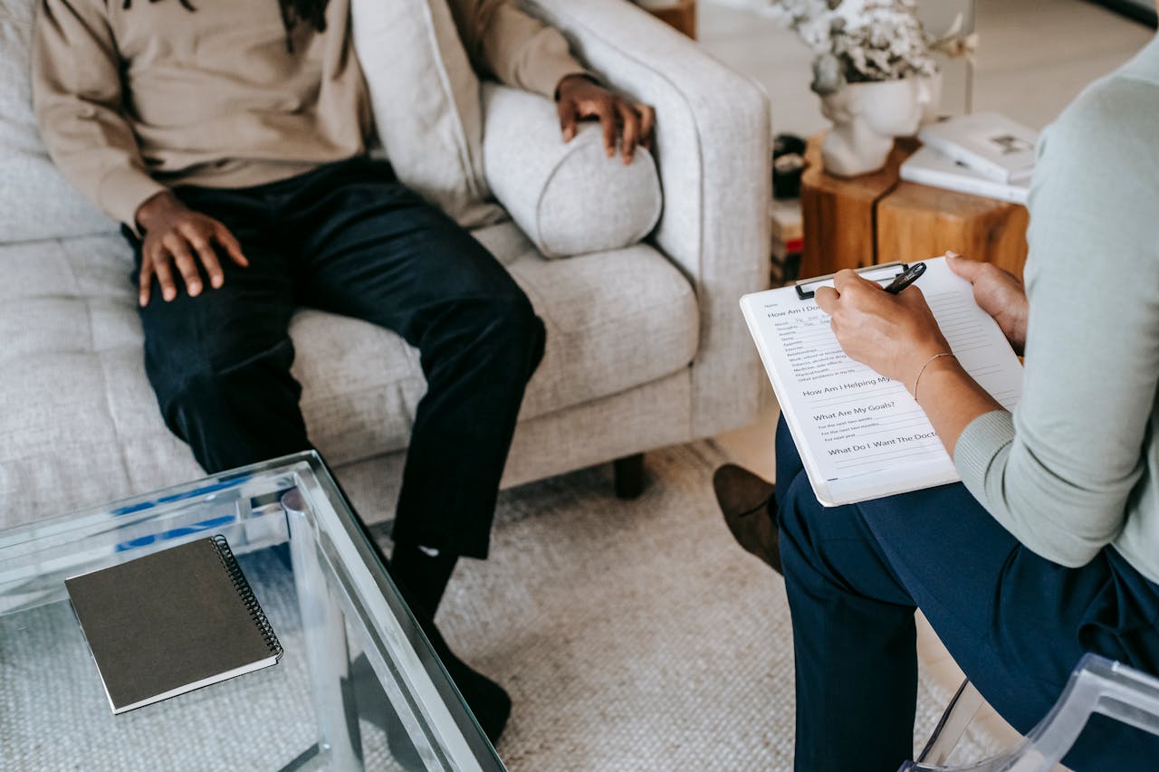 Two people sit in a living room; one holds a notebook and pen.