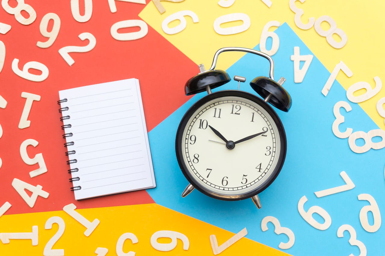 Black analog clock set to 10:10 on a colorful background with scattered wooden numbers and an open notebook.