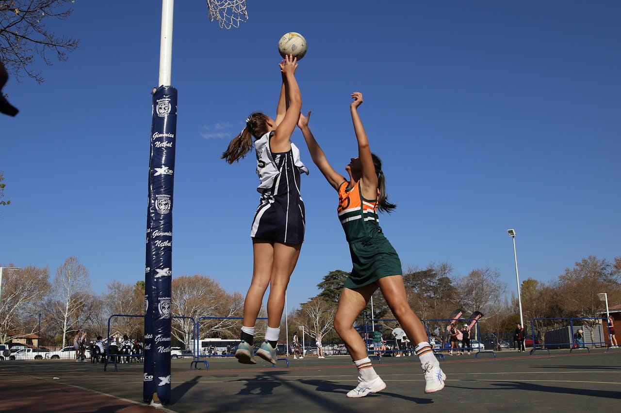 Two netball players jump to reach the ball near the hoop during an outdoor game on a sunny day.