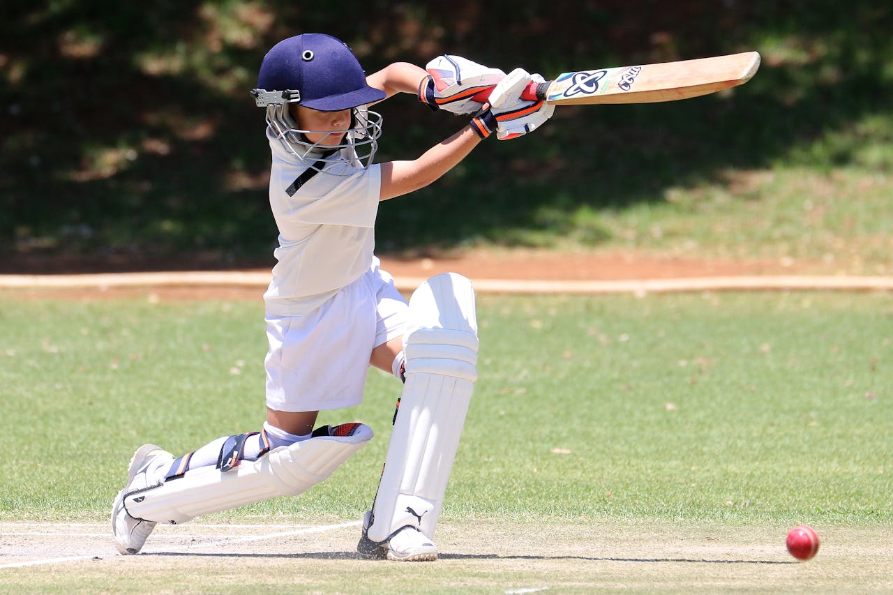 Cricket Young cricket player in white uniform and navy helmet prepares to hit a red ball on a grassy field.