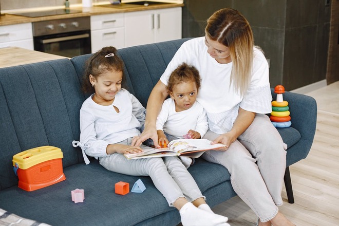 Children An adult and two children sit on a blue couch reading a book together. Toys are scattered around, and a kitchen with white cabinets is visible in the background.
