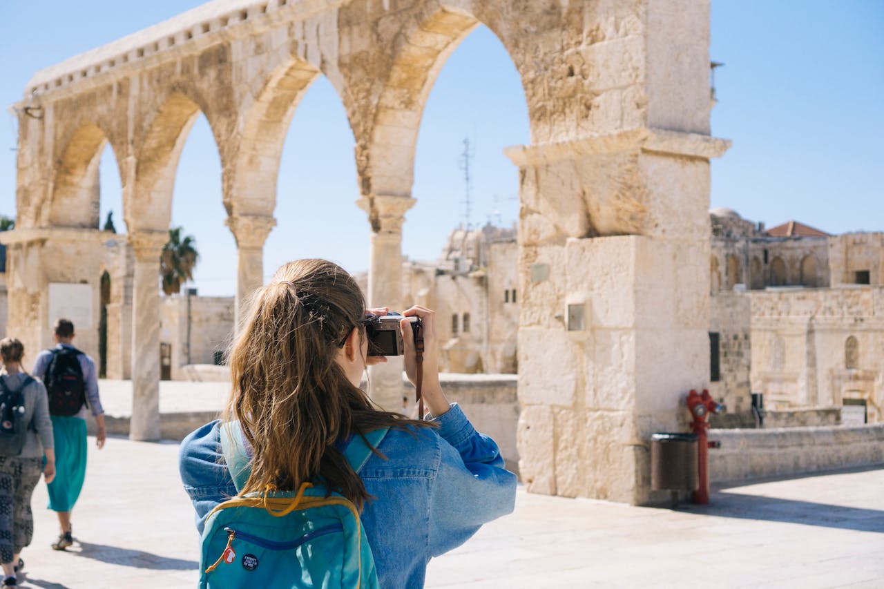 Person with a camera and backpack taking a photo near ancient stone arches under a clear sky.