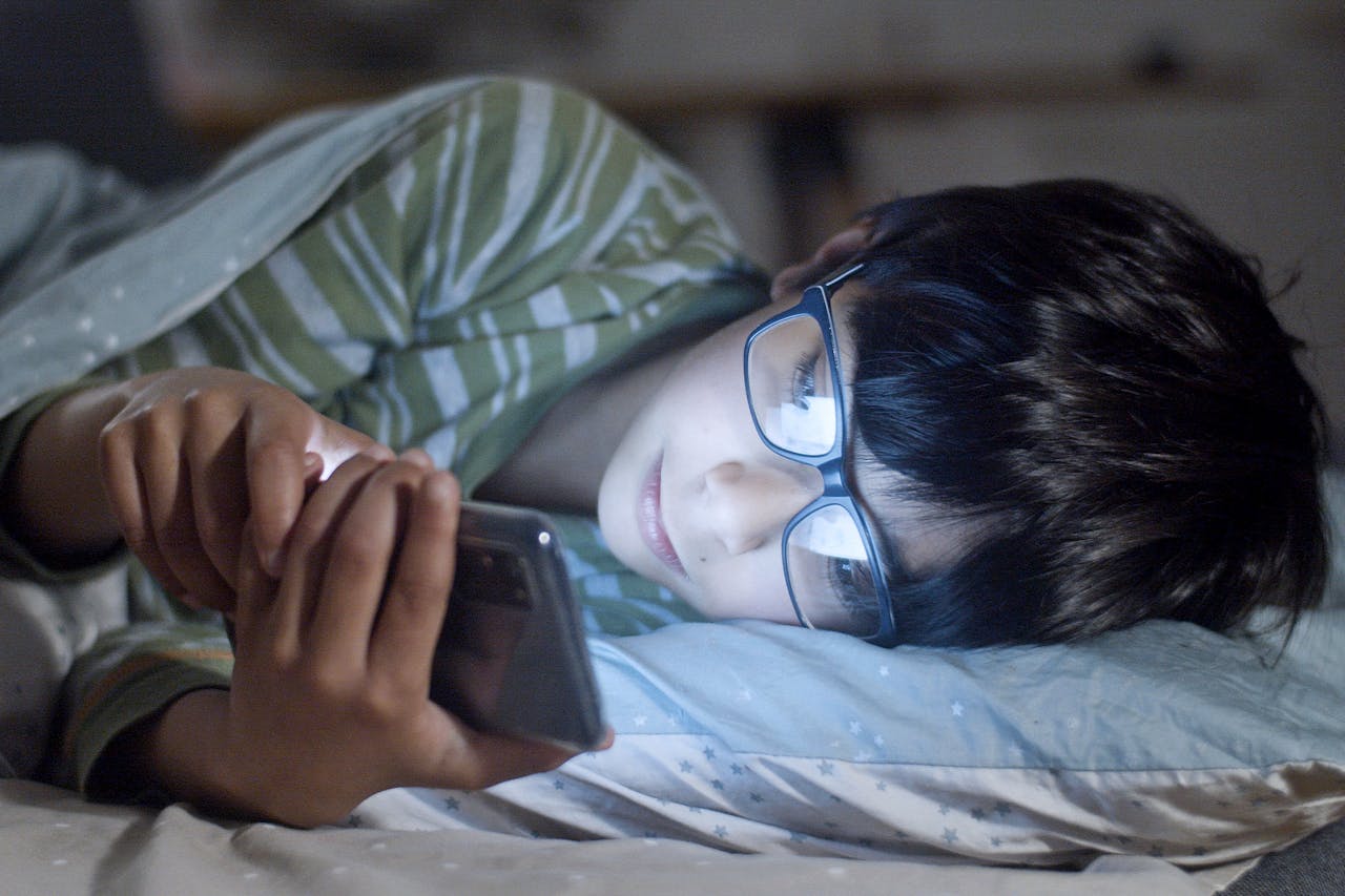 A boy is using a phone in bed before sleeping and his face is lit up by the glow of the phone screen
