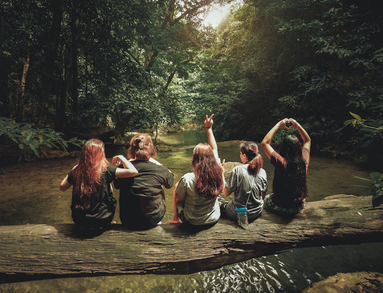Five women sit on a log in a river. They are wearing hiking gear. Their backs are to the camera.