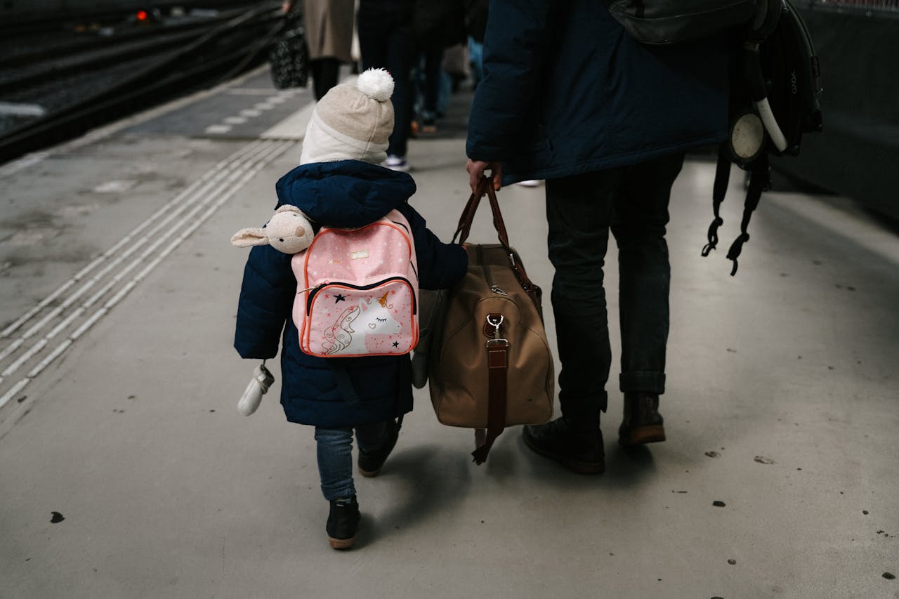 Child in blue coat with pink backpack and adult carrying bags walking on a train station platform.