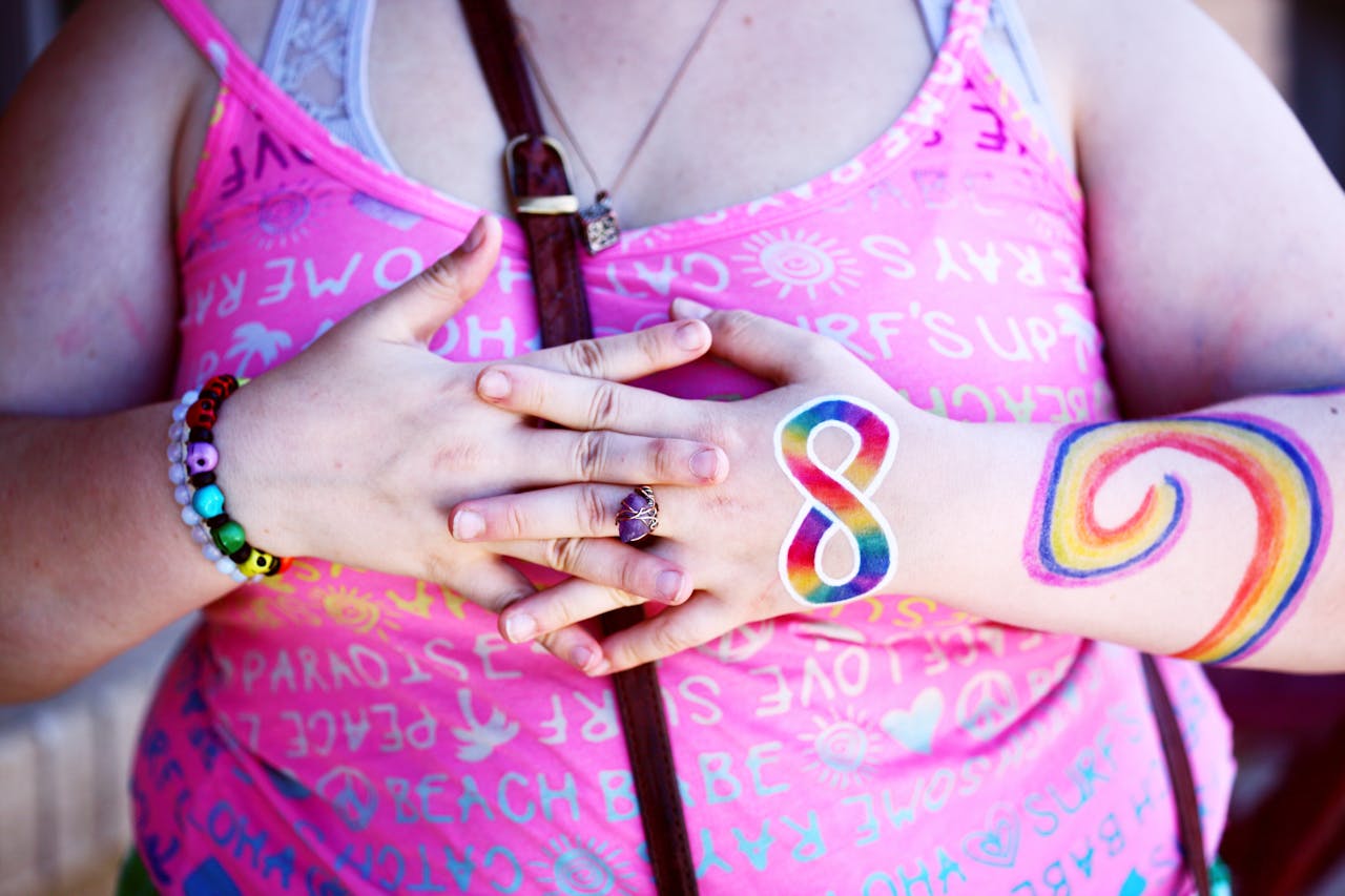 Person in a pink tank top with rainbow-painted arms, wearing bracelets and rings, hands clasped in front.