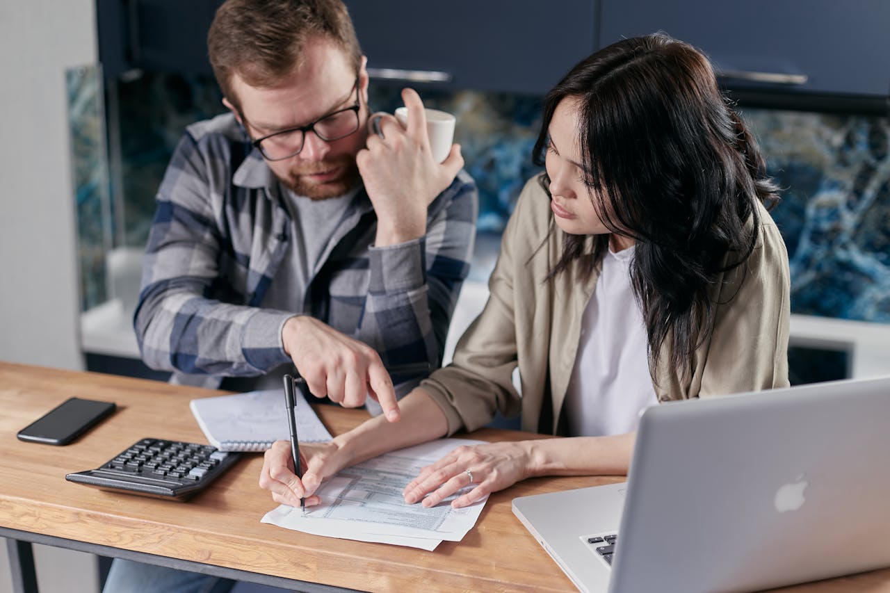 A man and a woman sit at a desk writing on a sheet of paper. They have a laptop and a calculator open.