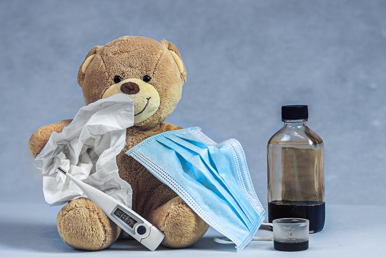 A teddy bear holding a tissue, thermometer and face mask is sat in front of a grey background. There is a bottle of medicine next to the teddy.
