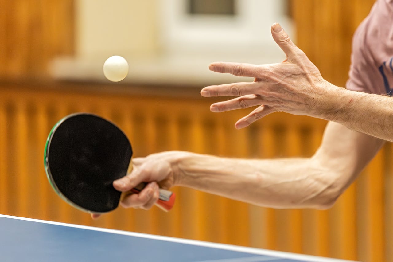 Table tennis Close-up of a person playing table tennis, holding a black paddle with a white ball mid-air.