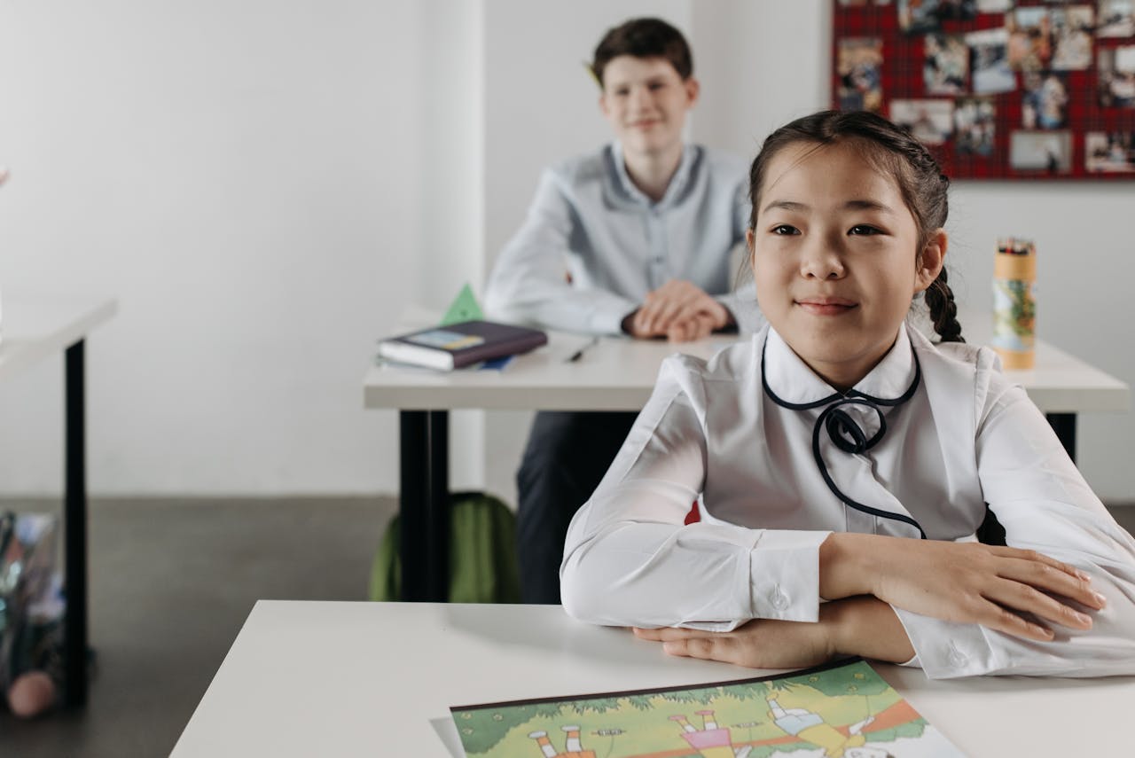 Two students seated at desks in a classroom with books and a bulletin board in the background.