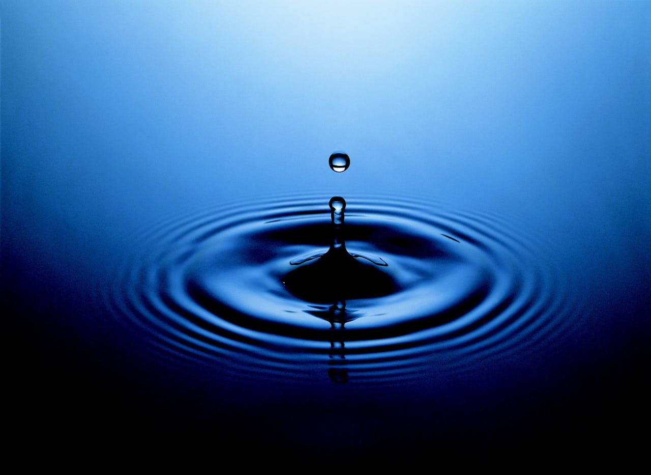 A water droplet suspended above ripples on a calm, blue surface.
