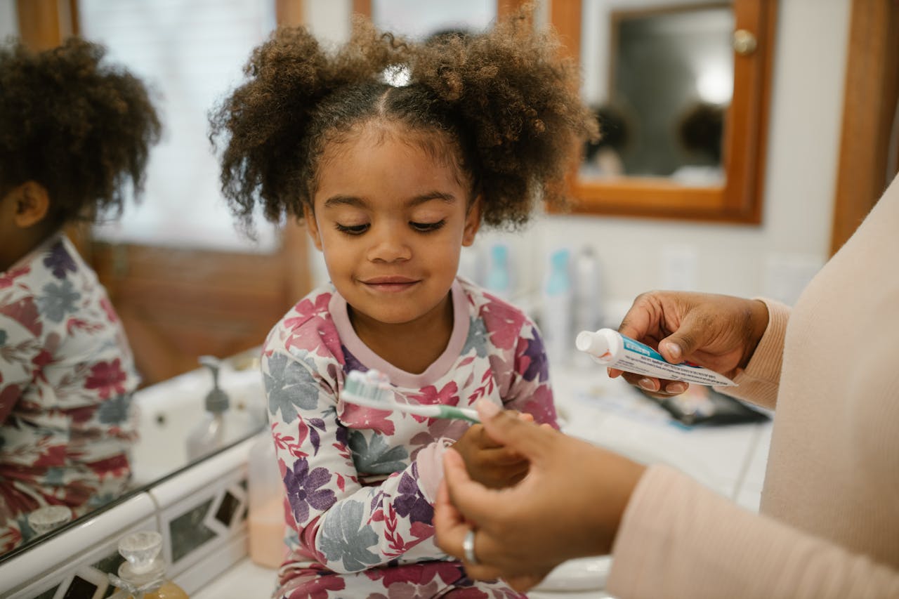 Child sitting on bathroom counter while an adult prepares a toothbrush with toothpaste; background includes bottles and a mirror.