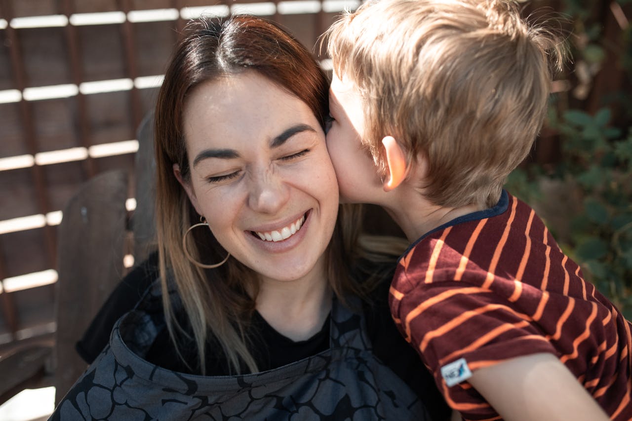 Child in a striped shirt kissing an adult on the cheek outdoors, with a wooden fence and greenery in the background.