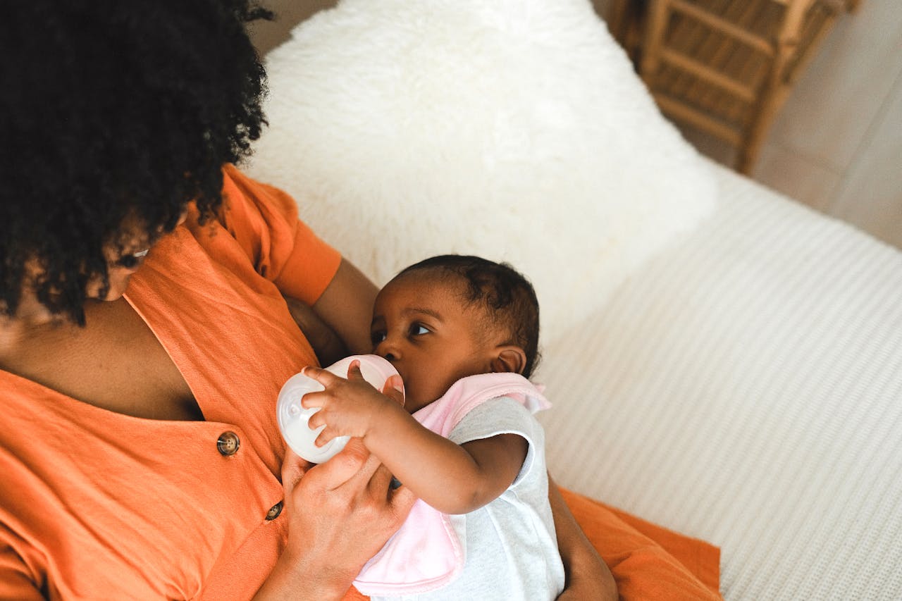 Bottle feeding A person in an orange shirt holds a baby wrapped in a white and pink blanket. The baby is drinking from a bottle. In the background, there is a white pillow.
