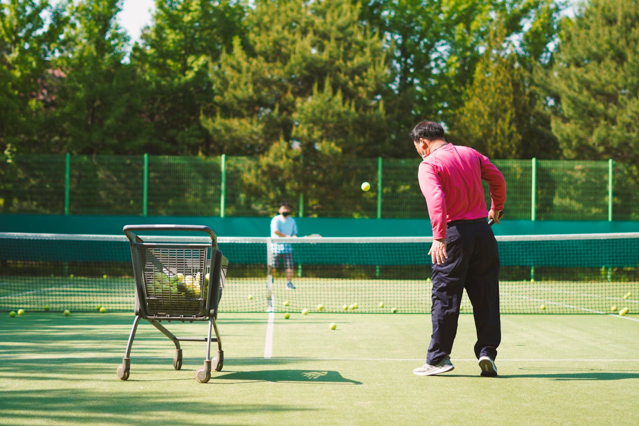 Playing tennis "Tennis coach in red shirt on court with ball basket, watching a player preparing to hit.