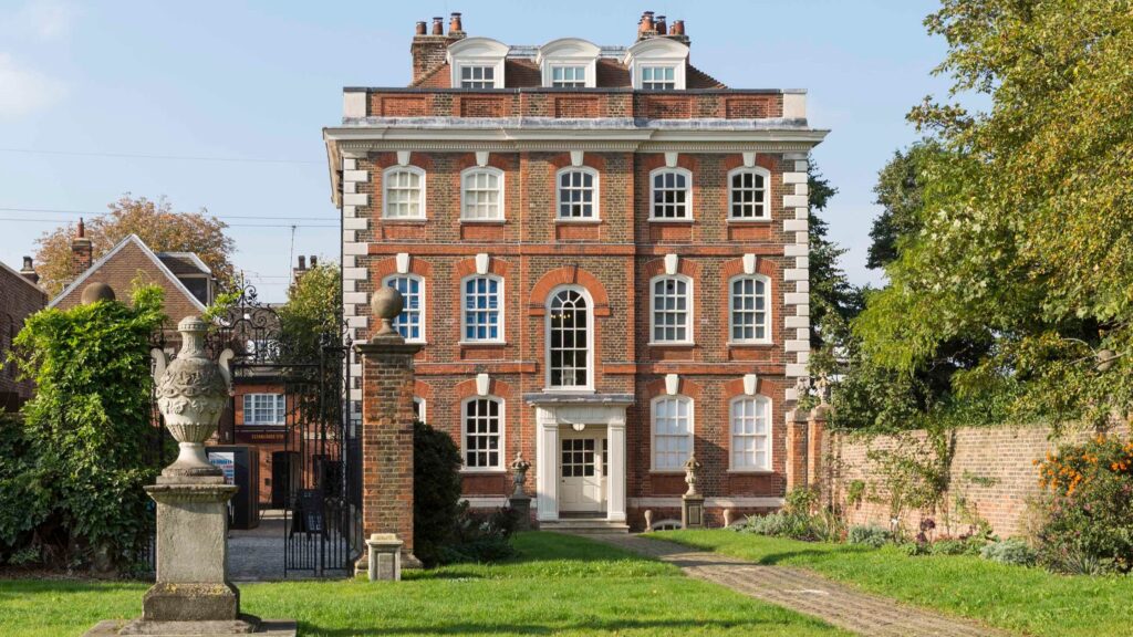 Four-story brick building with white window frames, central entrance, stone pathway, decorative pillars, and surrounding greenery.