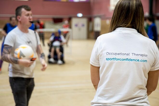 Person in a white 'Occupational Therapist' shirt standing in a gym; others, including someone in a wheelchair, are visible.