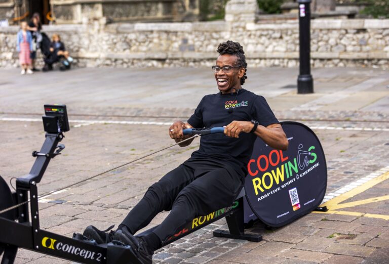 A person using a Concept2 rowing machine outdoors, wearing a black t-shirt with the words "COOL ROWING" and "ADAPTIVE SPORTS & EXERCISES."
