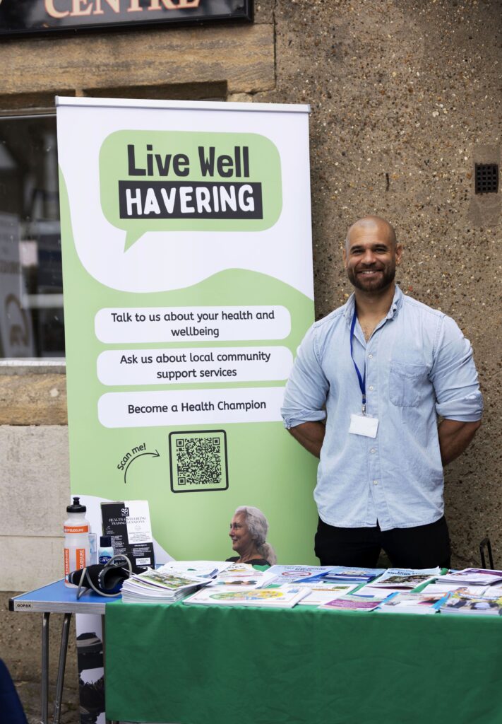 A Live Well Havering Health Trainer smiles in front of the Live Well Havering banner. He is standing at a green stall and is smiling.