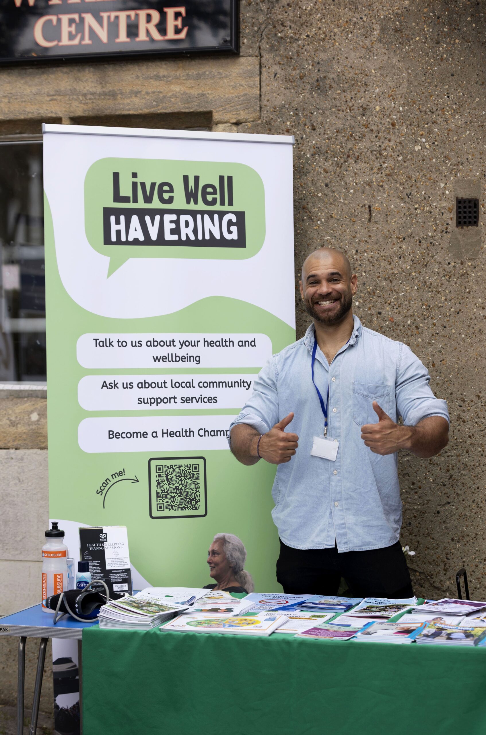 A Live Well Havering Health Trainer smiles in front of the Live Well Havering banner. He is standing at a green stall and is smiling with his thumbs up.