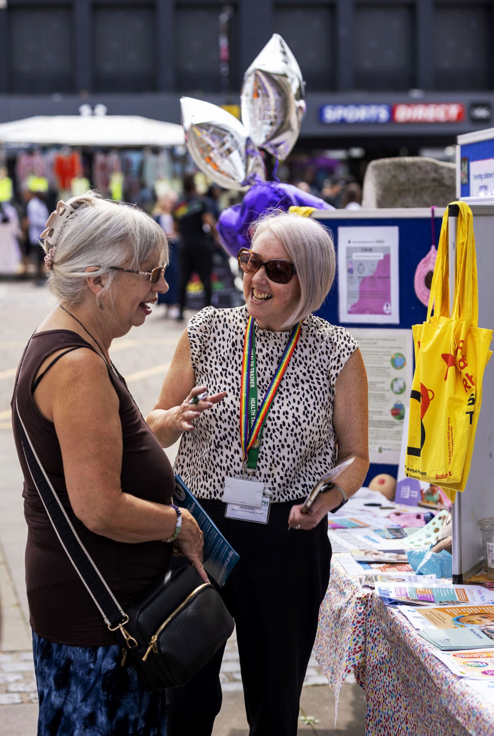 Two women stand at an outdoor information stall, smiling and chatting. One woman is wearing sunglasses, a rainbow lanyard, and a sleeveless patterned top, while the other carries a handbag and holds a phone. The table is covered with leaflets and health-related materials, with a yellow tote bag and posters displayed in the background.