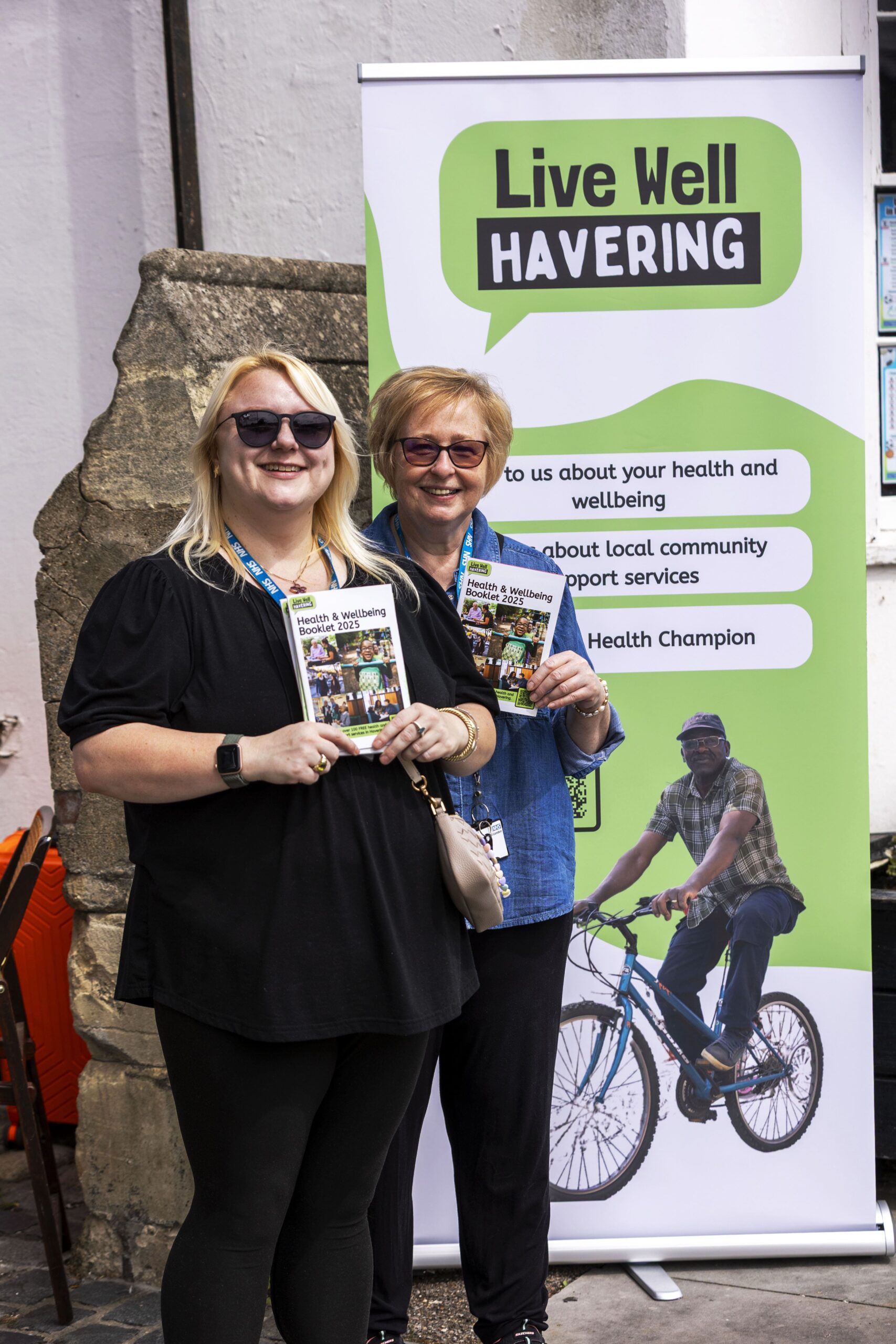 Two women stand smiling in front of a Live Well Havering banner, each holding informational leaflets. The banner promotes health, wellbeing, and community support services.