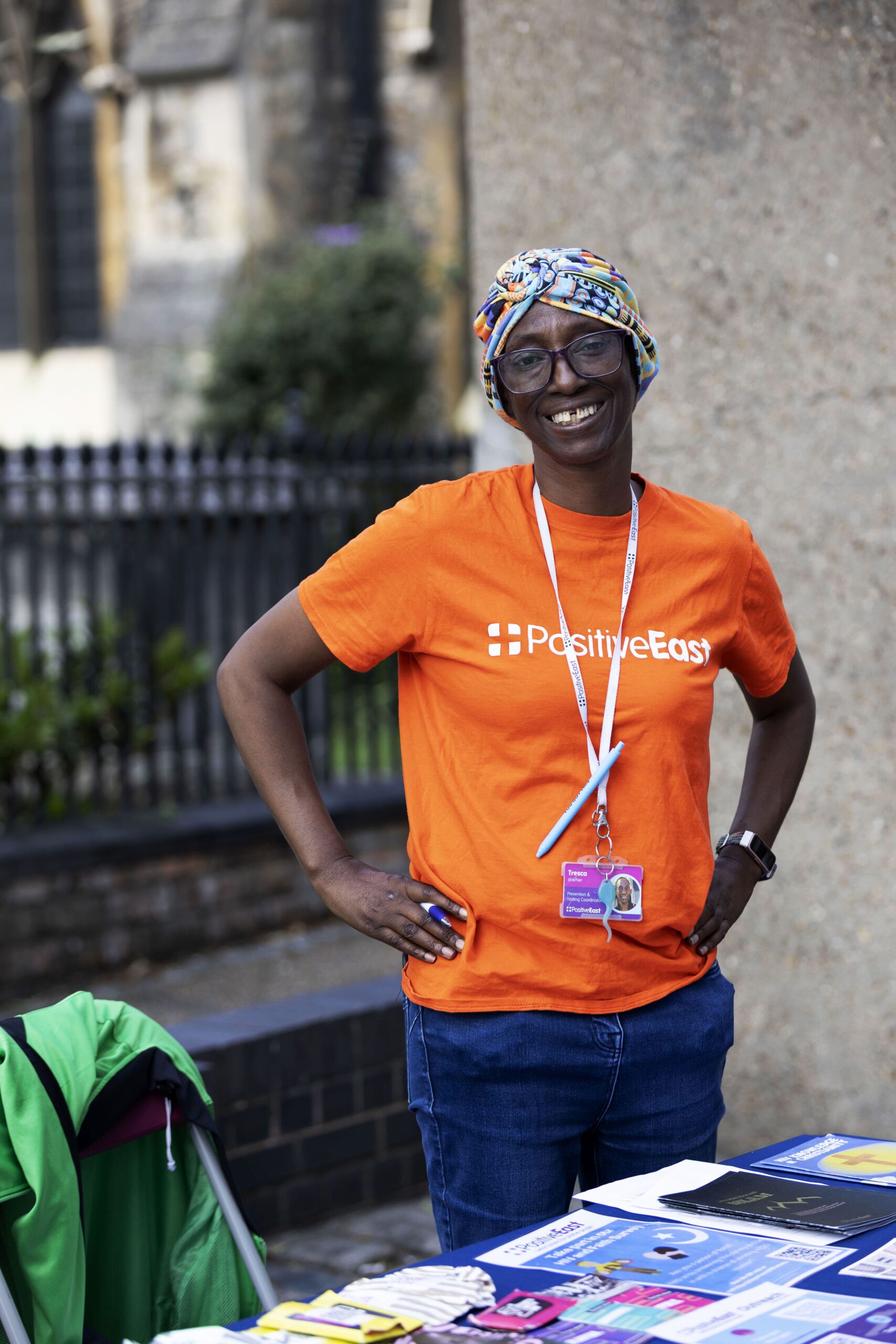 A smiling person in an orange Positive East t-shirt stands by an outdoor table with leaflets. They wear glasses, a colorful headscarf, and a lanyard with an ID badge. A building and iron fence are in the background.