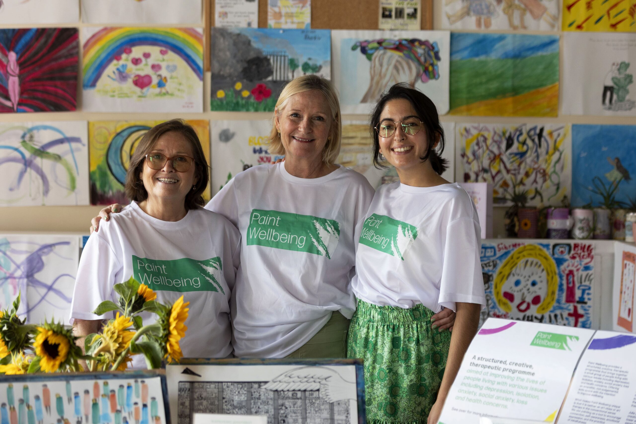 Three women wearing “Paint Wellbeing” shirts stand smiling in front of a colorful display of artwork. Sunflowers and informational boards are on the table in front of them. The background is filled with children’s drawings.
