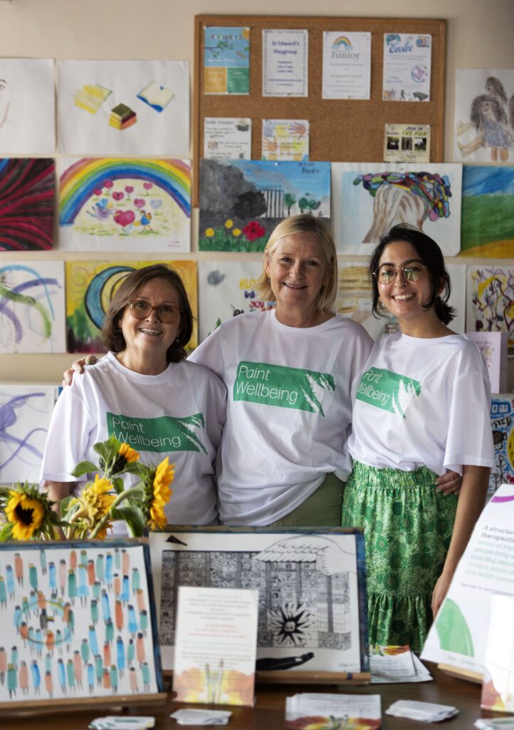 Three women wearing “Paint Wellbeing” shirts stand smiling in front of a display of colorful artwork and informational materials. Sunflowers and flyers are on the table in front of them.