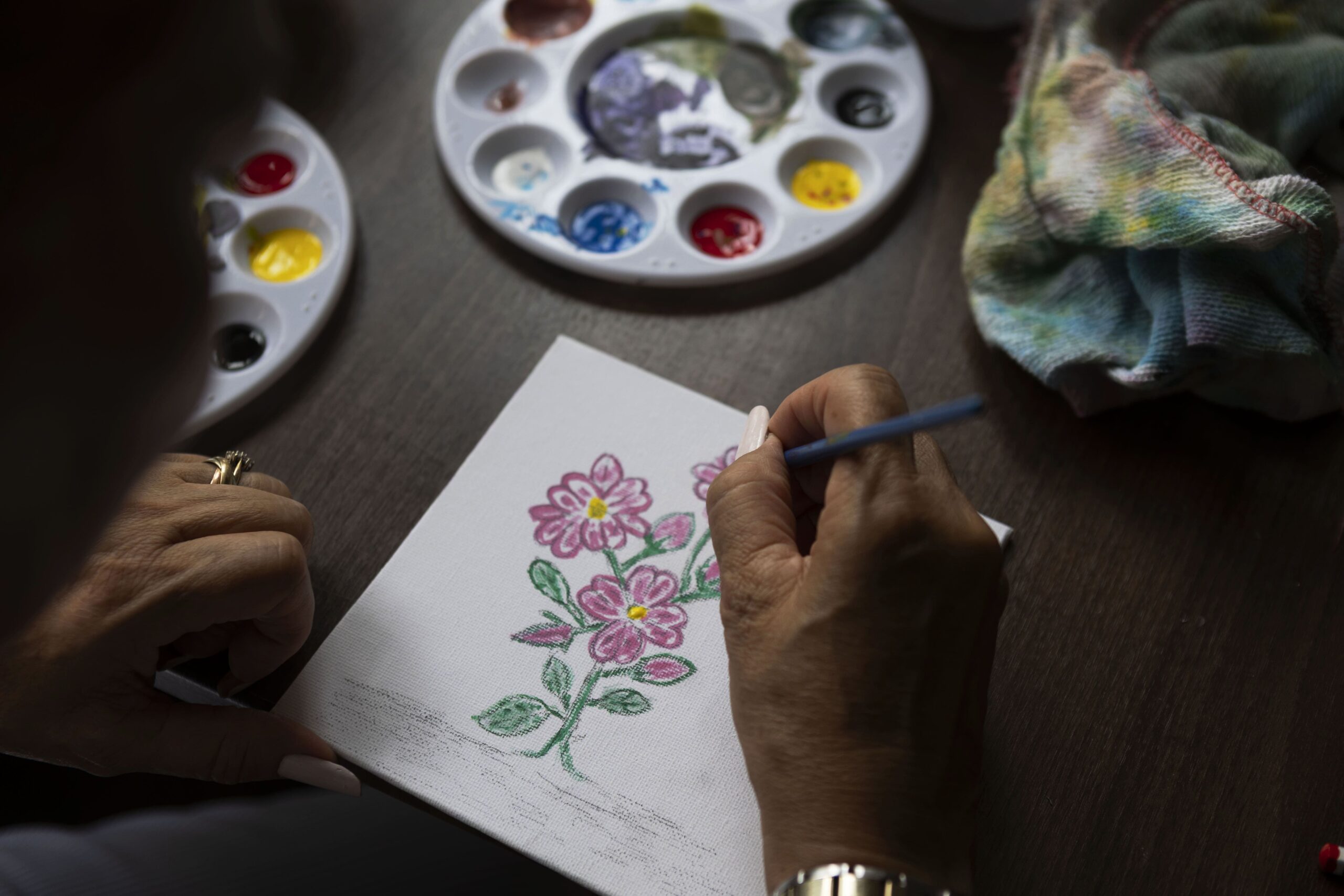 A person paints pink flowers with green stems on a small canvas, using a thin brush. Two paint palettes with various colors and a colorful cloth are on the wooden table nearby.