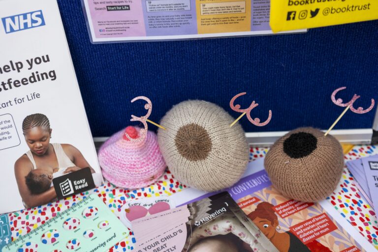 Three knitted breasts, used as breastfeeding teaching aids, are displayed on a table with informational leaflets and NHS breastfeeding support materials. The tablecloth is colorful and spotty.