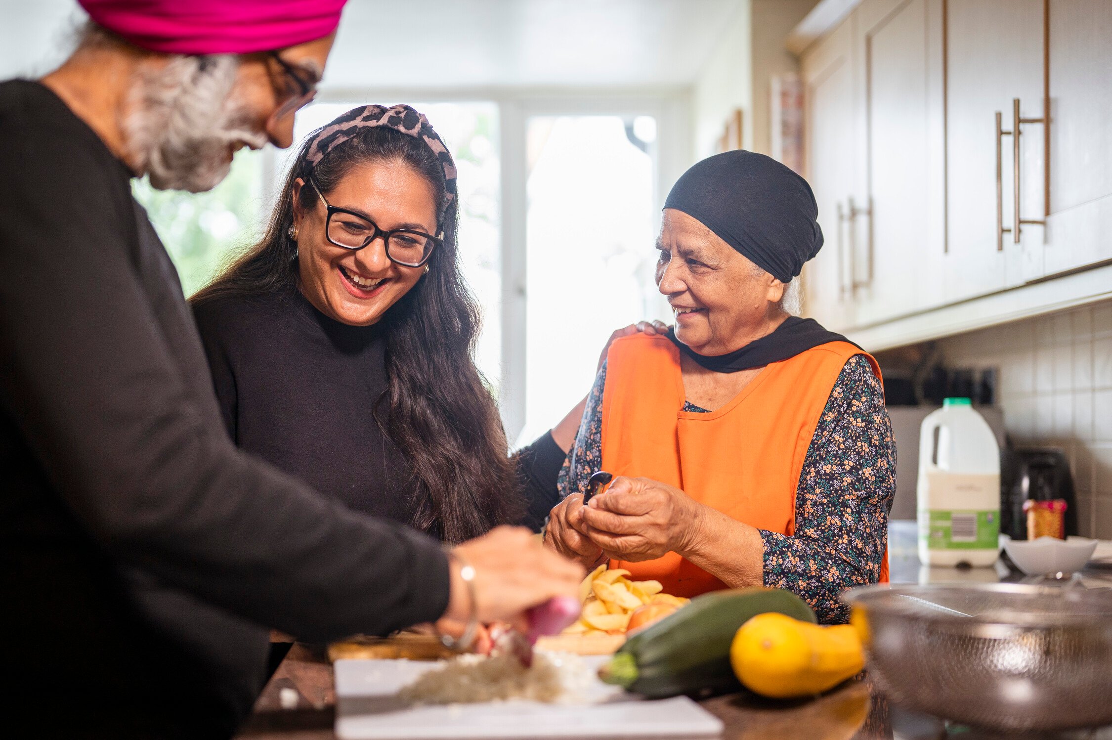Three people in a kitchen prepare food together, smiling and chatting. An older woman wears an orange apron, while a younger woman and a man in a pink turban are also helping with the meal prep. Vegetables are on the counter.
