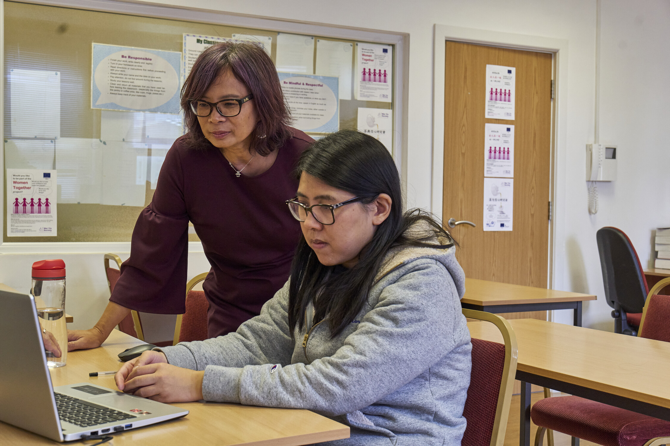 Two women in a classroom setting focus on a laptop. One is seated and using the laptop, while the other stands beside her, offering guidance. Papers and notices are visible on the walls in the background.