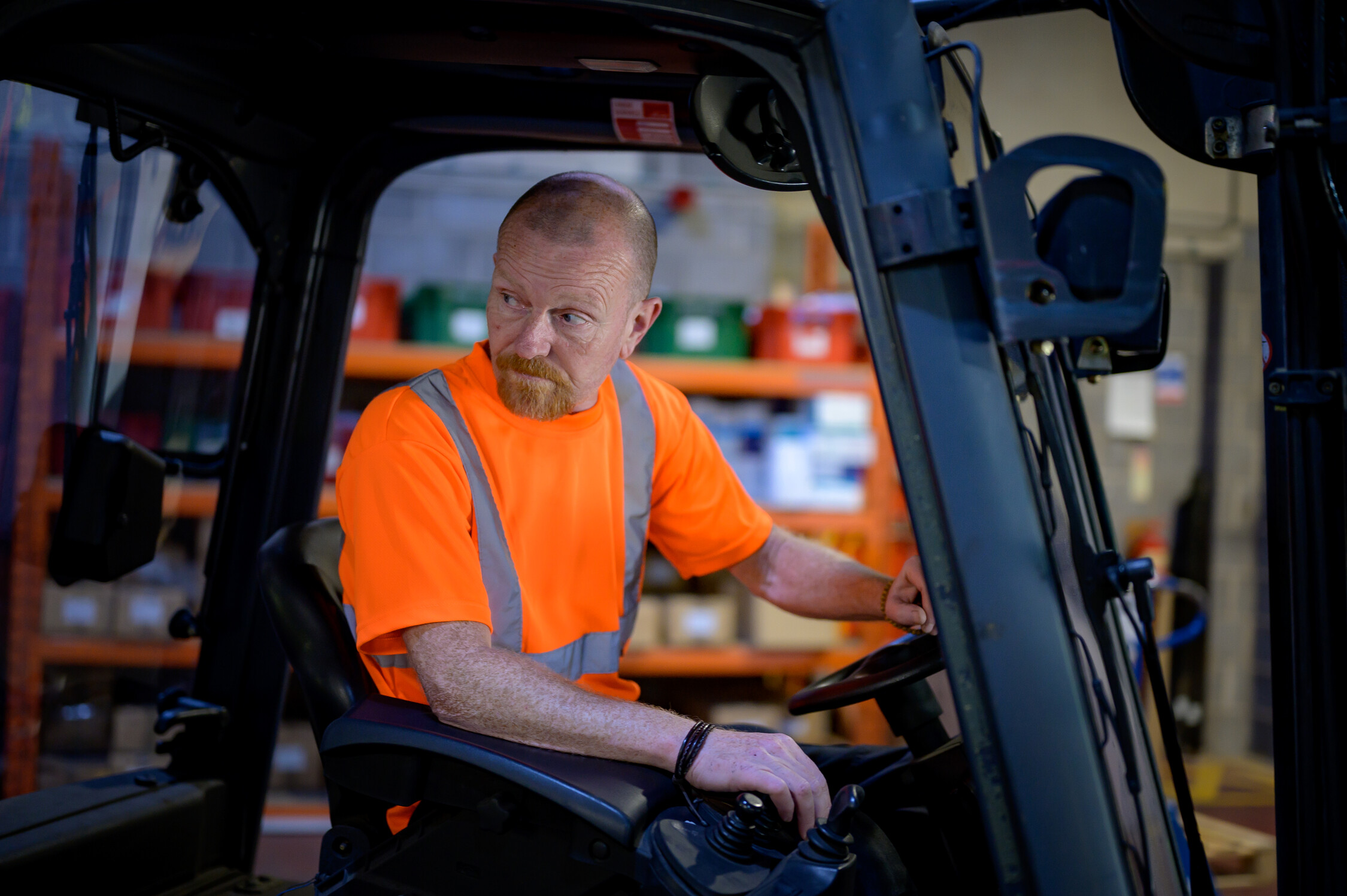 A bald man in orange high vis clothing reverses a forklift in a warehouse.