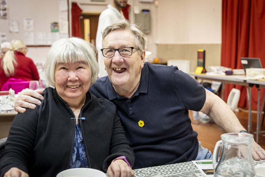 Two older adults, a woman and a man, sit close together at a table, smiling happily at the camera. The man’s arm is around the woman’s shoulders. The setting appears to be a community or social event.