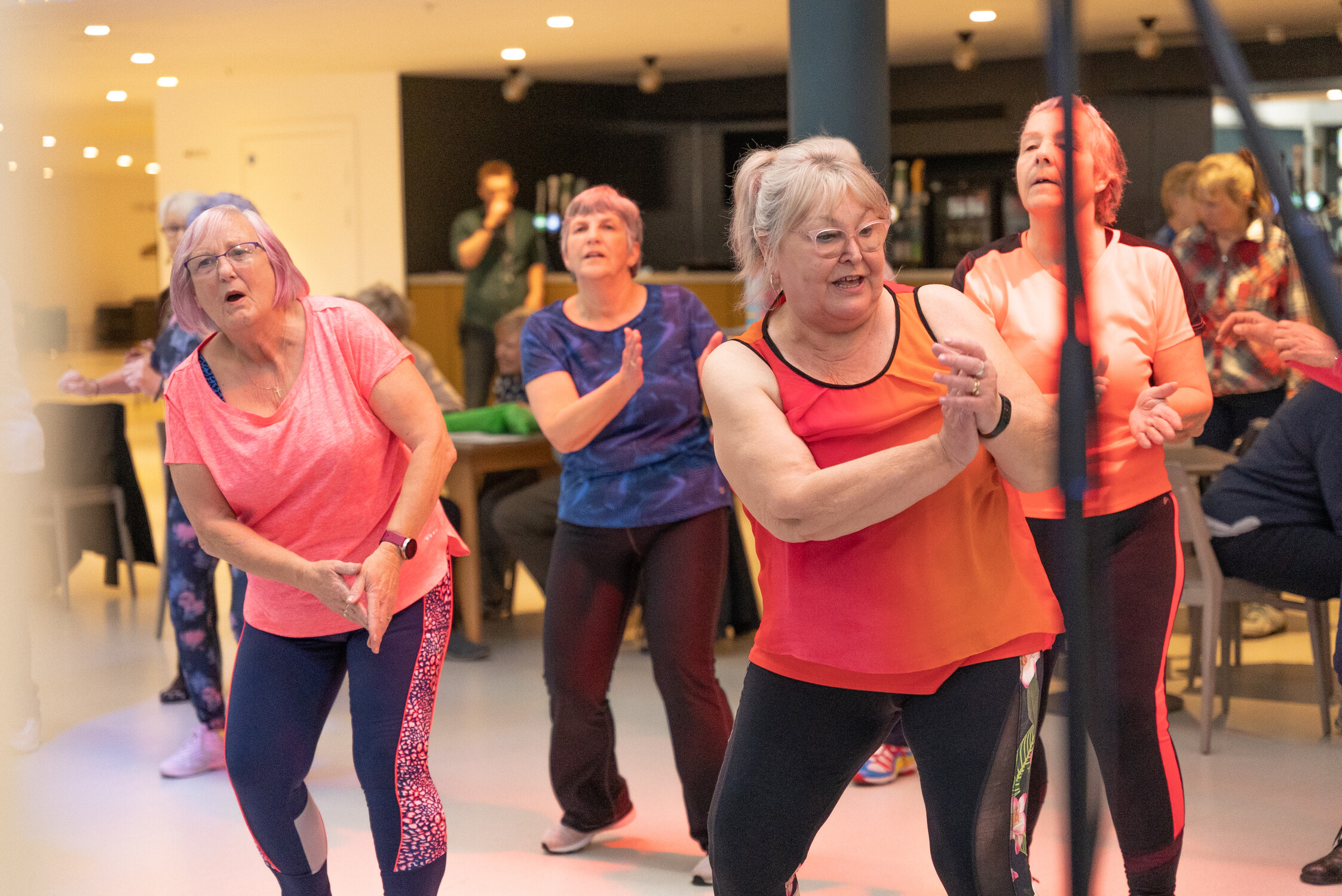 A group of older women in colorful workout clothes participate in a lively indoor dance or fitness class, moving energetically and smiling. Other people are visible in the background.