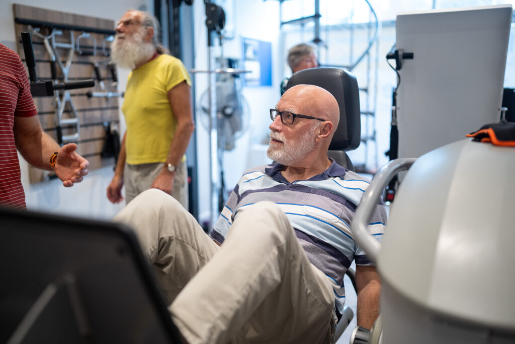 An older man with glasses uses a leg press machine at a gym, while another man in a yellow shirt and others are seen in the background. The setting appears bright and modern.