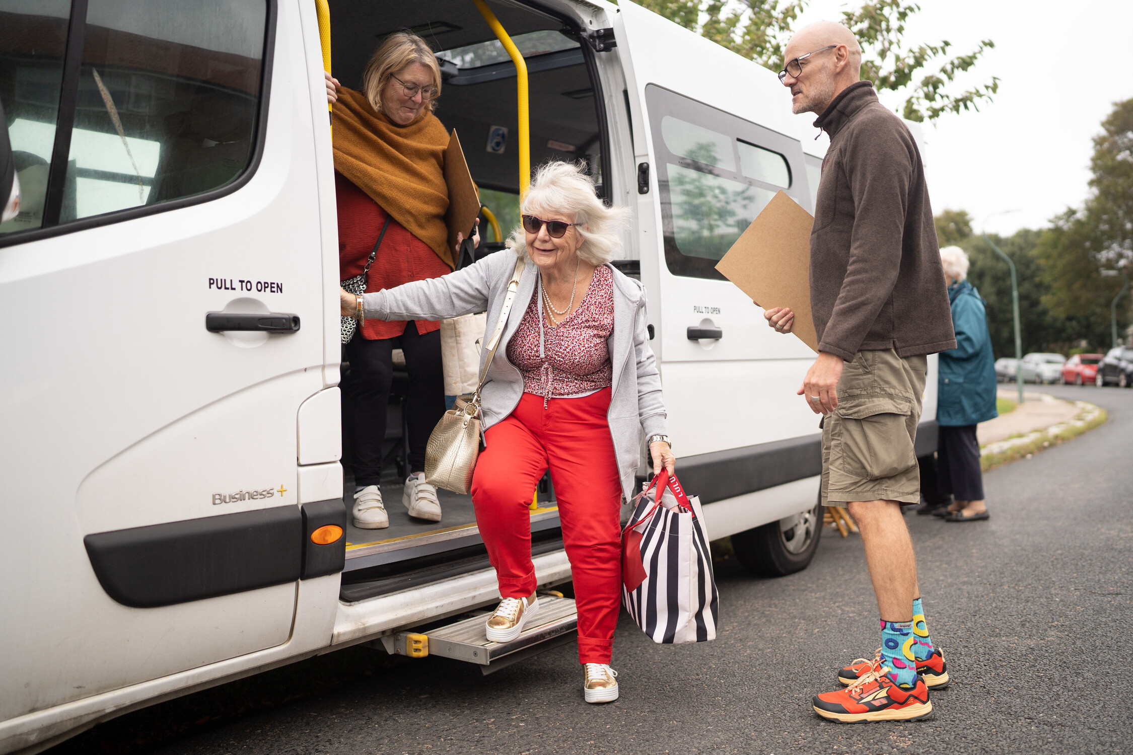 An older woman with white hair and sunglasses steps out of a white van holding a striped bag, as another woman follows behind her and a man in colorful socks stands nearby holding a clipboard.
