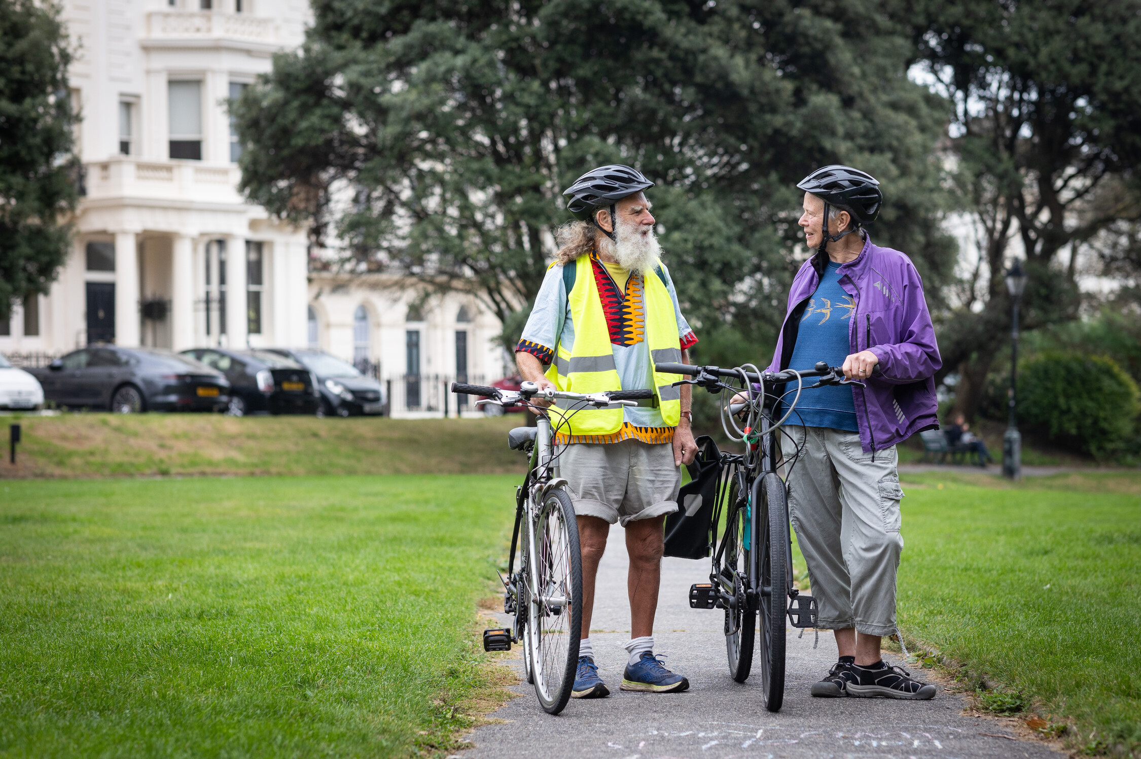 Two older adults wearing helmets and casual clothing stand with their bicycles on a park path, smiling at each other. Trees and white buildings are visible in the background.