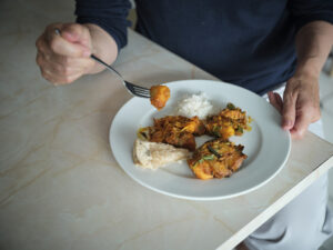A person holding a fork is about to eat from a white plate with rice, fish, and assorted vegetables or curry dishes, sitting at a light-colored table.